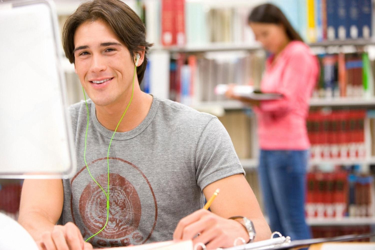 Smiling university student in a library preparing for an online exam with earphones and a laptop